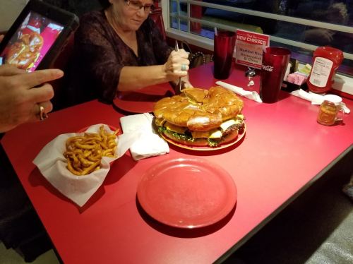 A woman sits at a red table, preparing to cut a large burger next to a plate of curly fries.