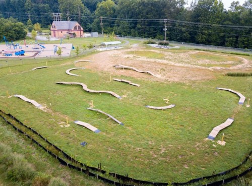 Aerial view of a grassy area with winding paths and wooden ramps, near a playground and surrounded by trees.