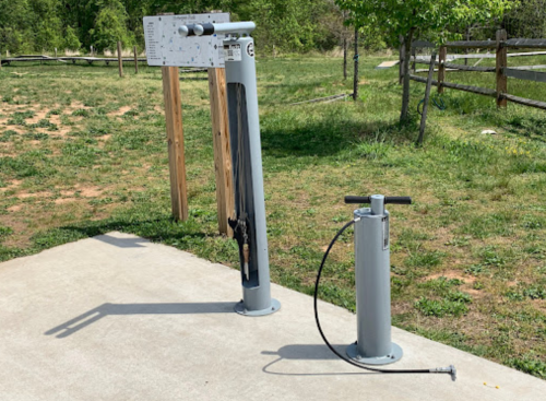 Outdoor fitness equipment featuring a vertical machine and a hand pump on a concrete surface, surrounded by greenery.