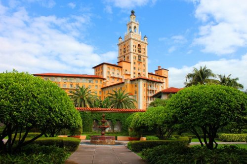 A grand orange hotel with a tall clock tower, surrounded by lush greenery and palm trees under a blue sky.