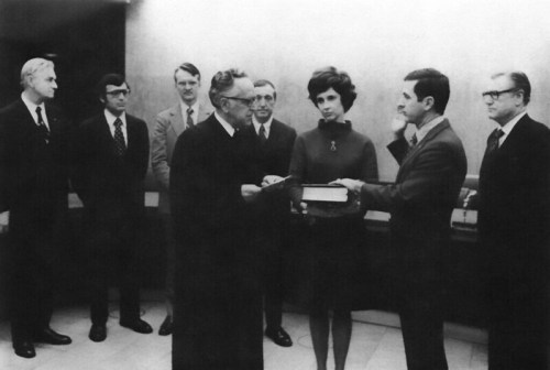 A black and white photo of a group of men and a woman in formal attire, gathered around a man holding a book.