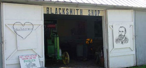 Exterior of a blacksmith shop with open doors, featuring a welcome sign and a portrait of a man on the wall.