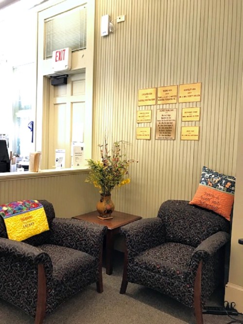 Two patterned chairs with colorful pillows are positioned around a small table, next to a wall with informational signs.