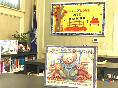 A cozy library scene featuring a "Reading to Children" display with teddy bears and a colorful fall-themed bulletin board.