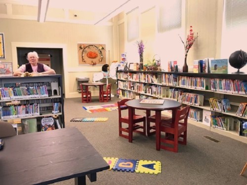 A cozy library with bookshelves, a globe, and a smiling person at a table surrounded by colorful rugs and children's books.