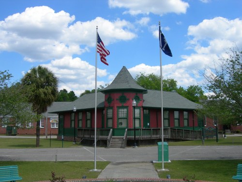A green and red building with a pointed roof, flanked by American and state flags, set against a blue sky with clouds.