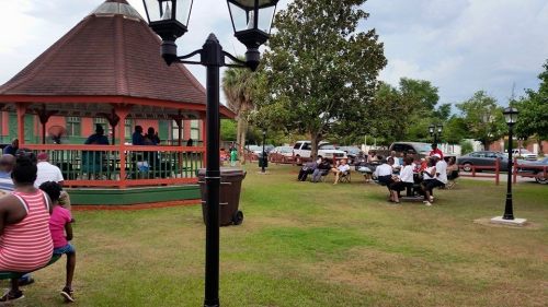 A park scene with a gazebo, people sitting on benches, and a few cars in the background under a cloudy sky.