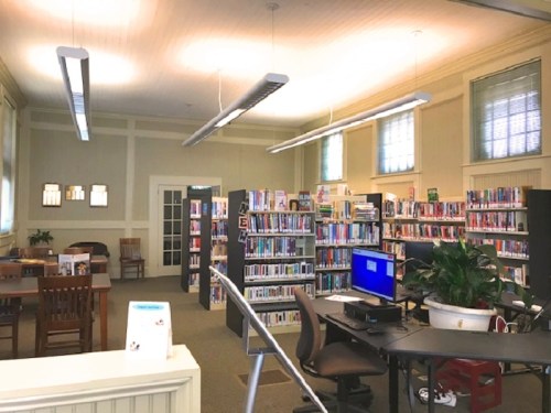 A spacious library interior with bookshelves, a computer station, and reading tables under bright overhead lights.