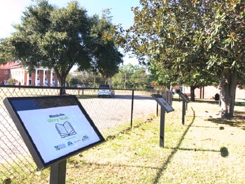 A fenced area with story walk signs and trees, set along a pathway near a building in a sunny outdoor setting.