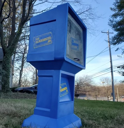 A blue, vintage-style newspaper box with a "Free Blockbuster" sign, set in a grassy area with trees in the background.