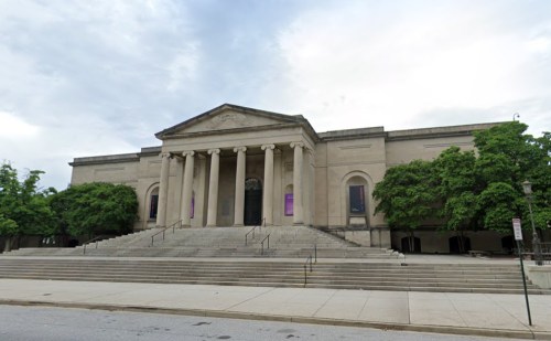 A large, classical-style building with columns and steps, surrounded by trees under a cloudy sky.