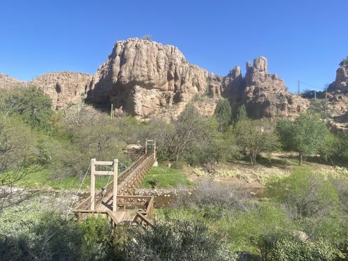 A wooden suspension bridge crosses a stream, surrounded by lush greenery and rocky cliffs under a clear blue sky.