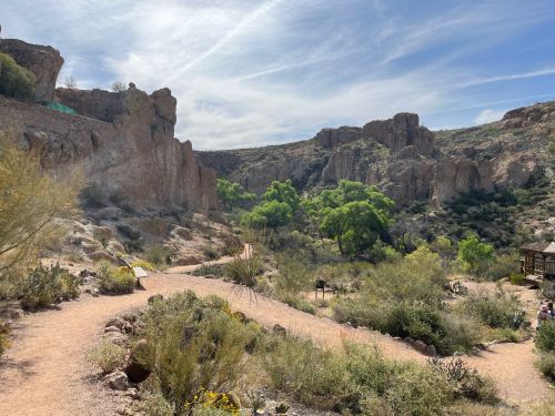 A scenic desert landscape with rocky cliffs, green trees, and a winding path under a blue sky with wispy clouds.
