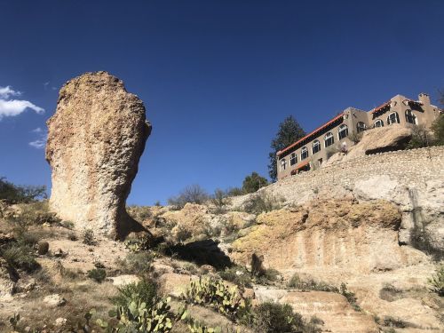 A large rock formation stands beside a multi-story building against a clear blue sky. Cacti are visible in the foreground.