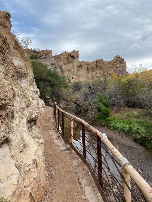 A narrow path along a creek, bordered by rocky cliffs and sparse vegetation under a cloudy sky.