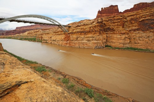 A curved bridge spans a river surrounded by rocky cliffs and desert vegetation under a cloudy sky.