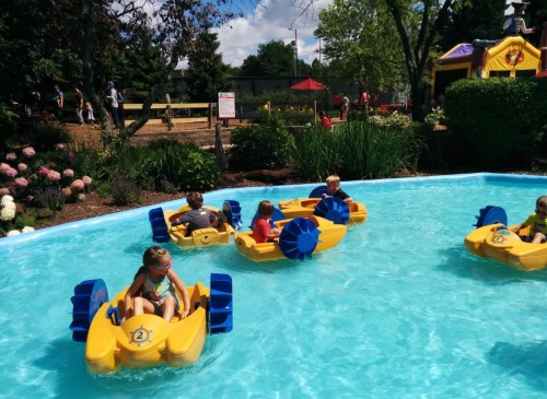 Children in colorful paddle boats enjoy a sunny day in a blue pool surrounded by greenery and flowers.