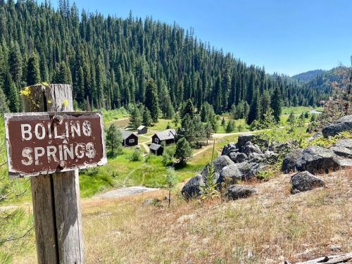 A wooden sign reading "Boiling Springs" in a forested area with a view of small cabins and mountains in the background.