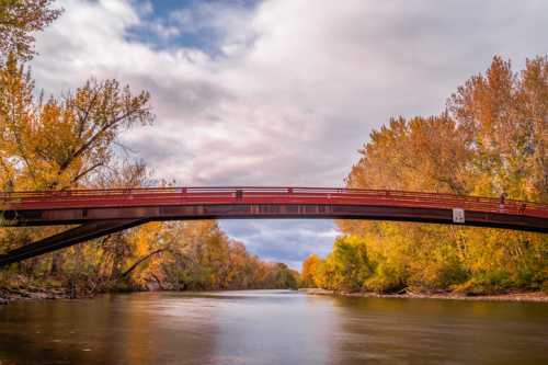 A red bridge spans a calm river, surrounded by trees with autumn foliage under a cloudy sky.