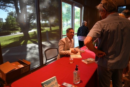 A man in a suit signs a book for a fan at a table, with large windows and greenery in the background.