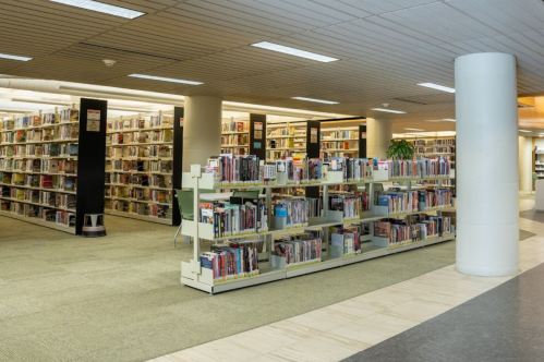 A spacious library interior with tall bookshelves and a central aisle, featuring rows of books and a few seating areas.
