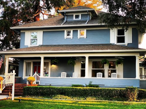 A charming blue two-story house with white trim, a front porch, and lush greenery in the yard.