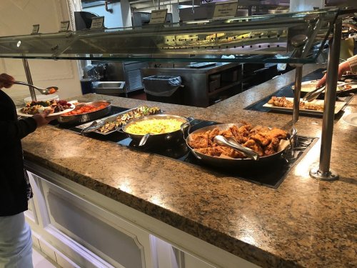 A buffet with various dishes, including fried chicken, pasta, and vegetables, under glass covers on a granite countertop.