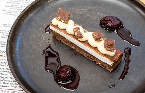 A slice of layered dessert with chocolate, cream, and caramel, garnished with chocolate sauce and berry compote on a plate.