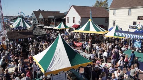 A bustling outdoor market with colorful tents, crowds of people, and shops along a waterfront street.