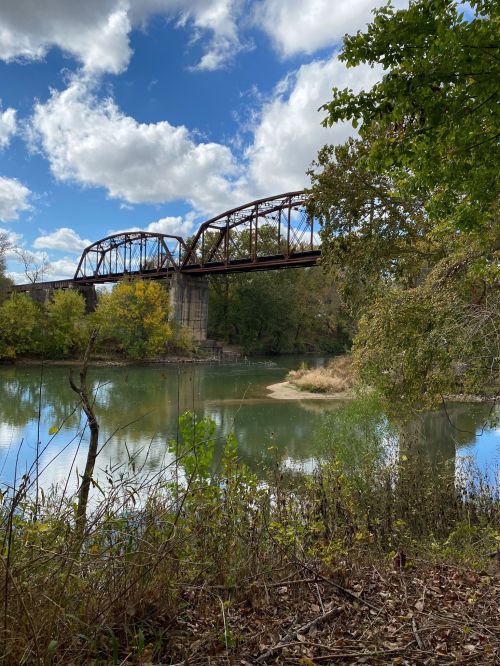 A rusted bridge spans a calm river, surrounded by trees and a blue sky with fluffy clouds.