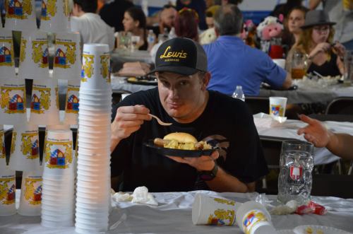 A man holds a plate of food, sitting at a crowded table with stacked cups and people in the background.