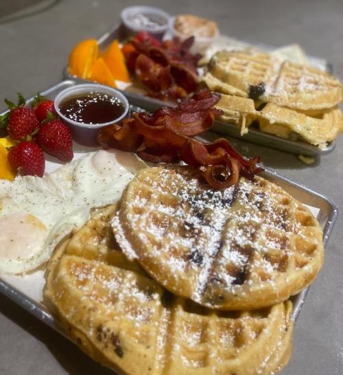 Two trays of breakfast featuring waffles, bacon, eggs, strawberries, oranges, and syrup.