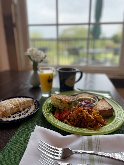 A breakfast spread featuring a sandwich, hash browns, fruit, a pastry, and a drink on a table with a scenic view.