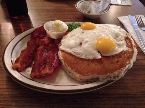 A plate of pancakes topped with two fried eggs, served with crispy bacon and a side of butter.