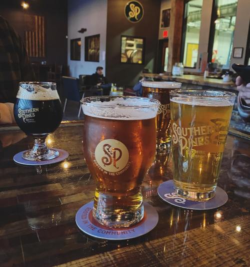 A wooden bar with three glasses of beer, each on coasters featuring the Southern Pines Brewing logo.