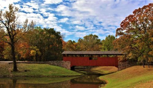 A red covered bridge spans a calm pond, surrounded by colorful autumn trees and a partly cloudy sky.