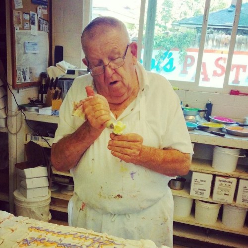 An elderly man decorates a cake with icing in a bright, busy kitchen filled with baking supplies.