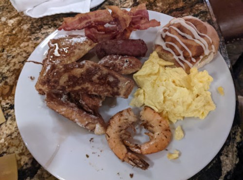 A plate of breakfast featuring French toast, bacon, sausage, scrambled eggs, shrimp, and a cinnamon roll.