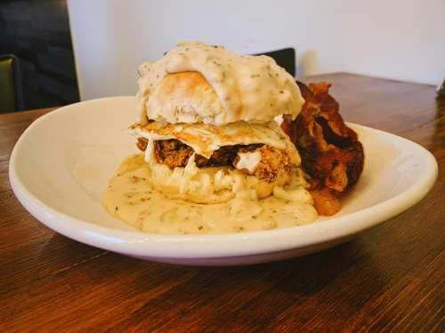 A biscuit sandwich with fried chicken, a fried egg, and creamy gravy, served with crispy bacon on the side.