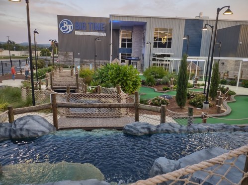 A mini-golf course with a water feature, surrounded by greenery, in front of a large building labeled "Big Time."