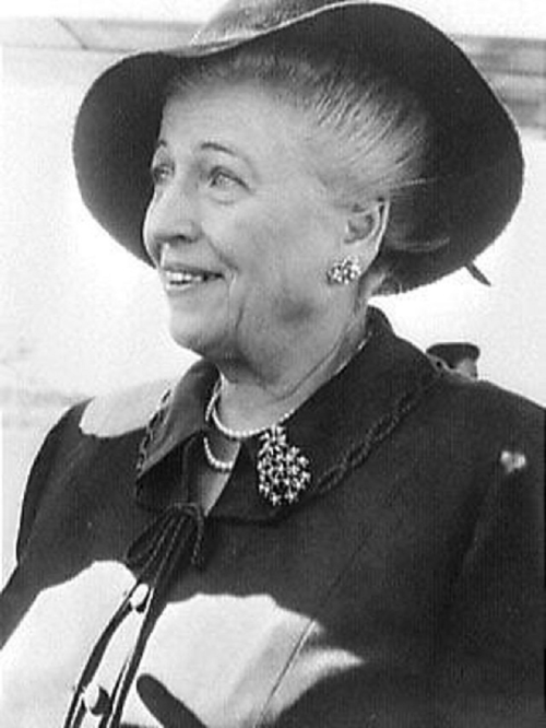 A smiling woman wearing a large hat and a stylish coat, adorned with a brooch, in a bright setting.