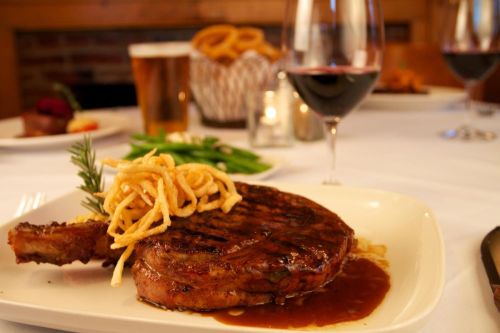 A plated steak with crispy onions, green beans, and red wine, set on a table with a beer and appetizers in the background.