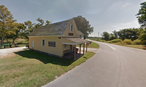 A yellow, two-story building at a roadside, surrounded by trees and open fields, with a sign and flags on the porch.