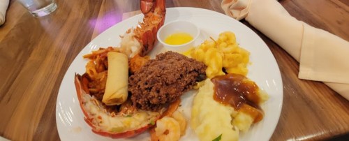 A plate featuring lobster, shrimp, macaroni and cheese, meatloaf, spring roll, mashed potatoes, and dipping sauce.