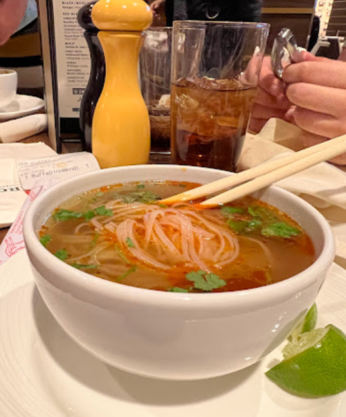 A bowl of soup with noodles and herbs, chopsticks resting on the edge, with a drink and a pepper shaker in the background.