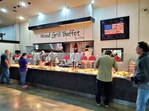 A buffet line with diners serving themselves, featuring a sign that reads "Wood Grill Buffet" and an American flag on the wall.