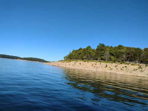 A calm lake reflects a clear blue sky, bordered by a tree-lined shore on a sunny day.