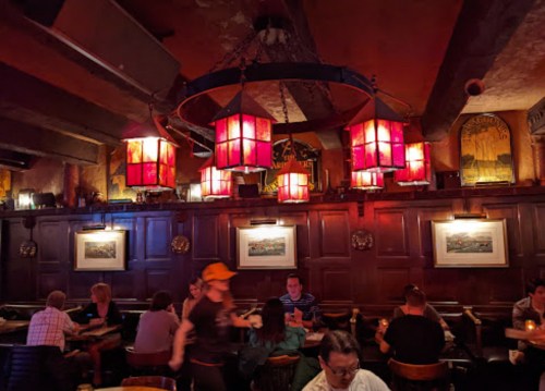 Cozy restaurant interior with warm lighting, wooden walls, and patrons dining at tables. Red lanterns hang from the ceiling.