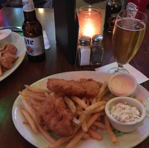 A plate of fried fish and fries, with coleslaw and dipping sauce, alongside a beer and a candle on a table.