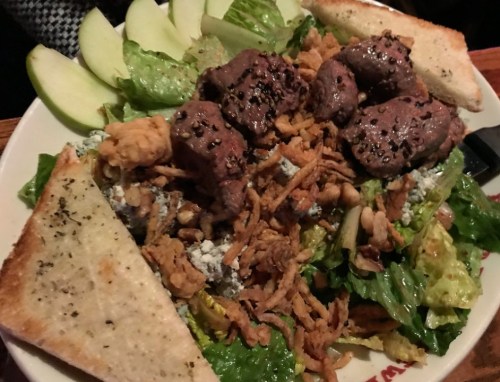 A hearty salad topped with grilled beef, crispy onions, lettuce, apple slices, and toasted bread on the side.